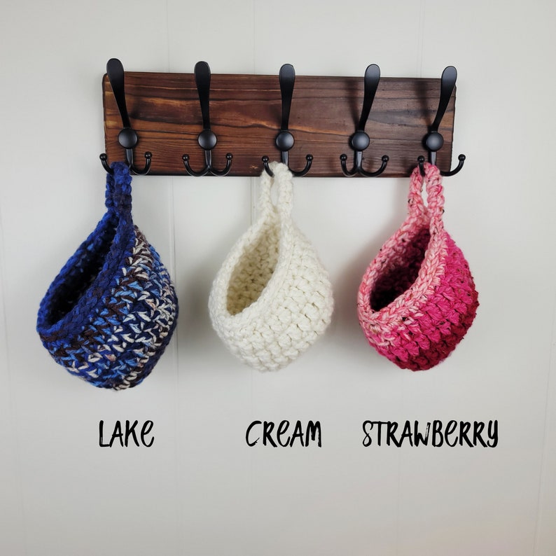 May include: Three crocheted hanging baskets in different colours: blue and brown, white, and pink. The baskets are hanging from hooks on a wooden rack. The baskets are labelled "Lake", "Cream", and "Strawberry".
