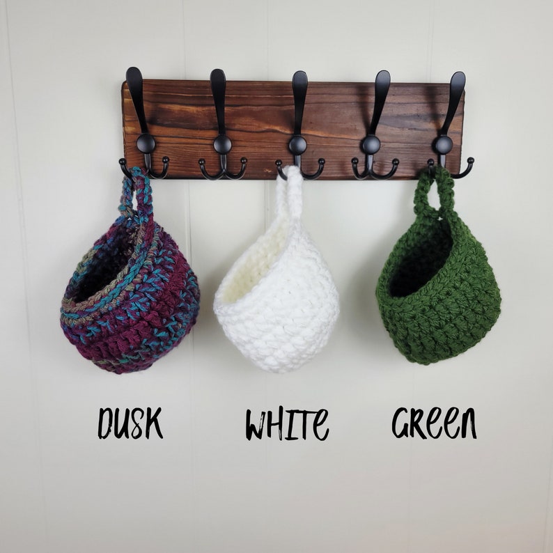 May include: Three crocheted hanging baskets in different colours: dusk, white, and green. The baskets are hanging from hooks on a wooden rack.