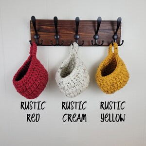 May include: Three hanging crocheted baskets in red, cream, and yellow colours. The baskets are hanging from hooks on a wooden wall rack. The text below the baskets reads "Rustic Red", "Rustic Cream", and "Rustic Yellow".