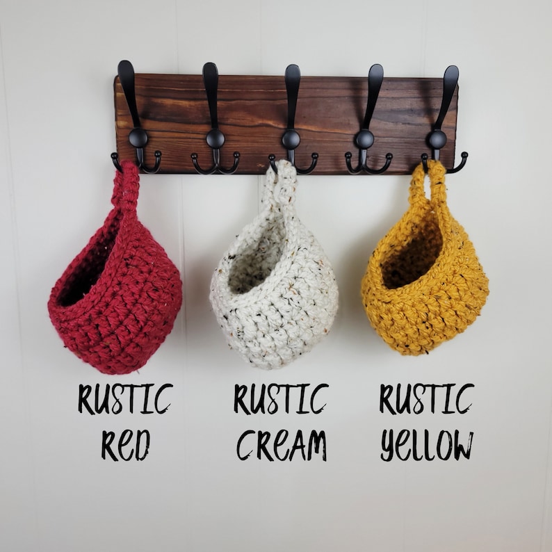 May include: Three hanging crocheted baskets in red, cream, and yellow colours. The baskets are hanging from hooks on a wooden wall rack. The text below the baskets reads "Rustic Red", "Rustic Cream", and "Rustic Yellow".