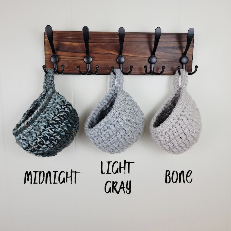 May include: Three hanging crocheted baskets in different colours: midnight, light grey, and bone. The baskets are hanging from a wooden rack with black hooks.