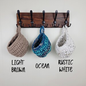 May include: Three hanging crocheted baskets in light brown, blue and white colours. The baskets are hanging from a wooden rack with black hooks. The text below the baskets reads "Light Brown", "Ocean" and "Rustic White".