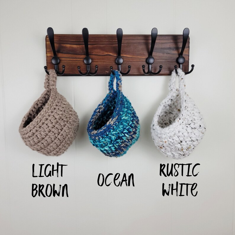 May include: Three hanging crocheted baskets in light brown, blue and white colours. The baskets are hanging from a wooden rack with black hooks. The text below the baskets reads "Light Brown", "Ocean" and "Rustic White".