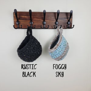 May include: A wooden wall mounted coat rack with six hooks. Two crocheted hanging baskets are on the hooks. One basket is black with white flecks and the other is blue, white, and brown. The text "Rustic Black" is below the black basket and "Foggy Sky" is below the blue basket.