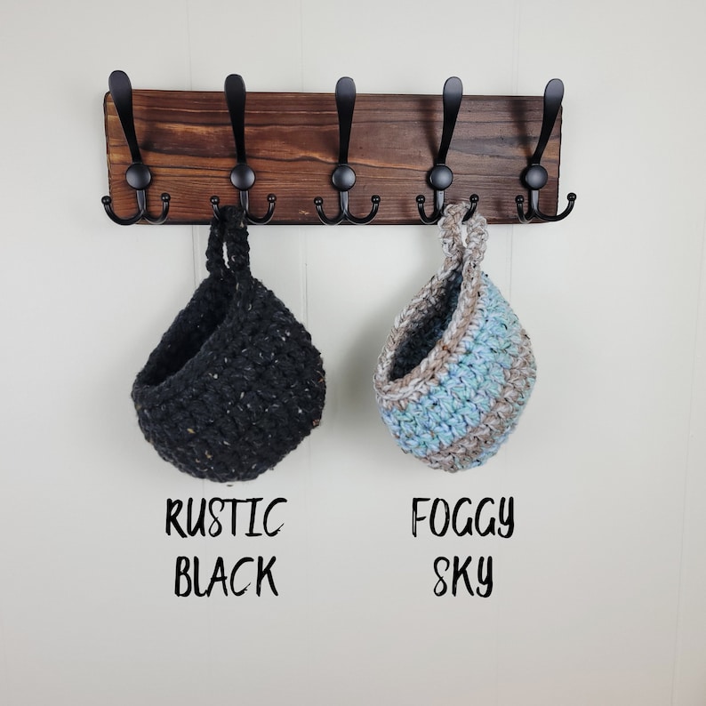 May include: A wooden wall mounted coat rack with six hooks. Two crocheted hanging baskets are on the hooks. One basket is black with white flecks and the other is blue, white, and brown. The text "Rustic Black" is below the black basket and "Foggy Sky" is below the blue basket.