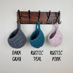 May include: Three crocheted hanging baskets in dark grey, teal, and pink, labelled "Dark Grey", "Rustic Teal", and "Rustic Pink", hanging from a wooden rack with black hooks.