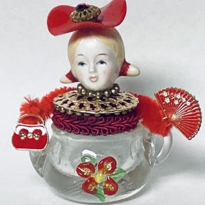 May include: A glass teapot figurine with a porcelain doll head wearing a red hat and a red and gold necklace. The teapot has a red flower design and a red purse attached to the handle.