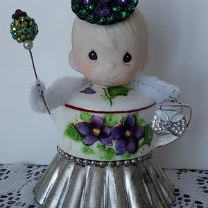May include: A Precious Moments figurine, featuring a doll-like figure with a floral-decorated teapot body and a silver skirt. The figure wears a jeweled hat and holds a wand and purse. The teapot is adorned with purple violets.