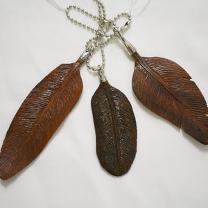 May include: Three brown leather feather charms on silver chains. The feathers are detailed with carved lines and have a rustic look.