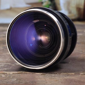 May include: A close-up of a camera lens with a black and silver finish. The lens has a reflective, purple-tinted glass. The lens is resting on a weathered, brown wooden surface. The background is blurred, showing a room with a hanging chair.