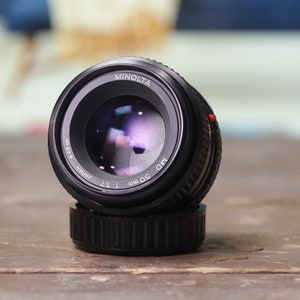 May include: A black Minolta MD 50mm f/1.7 lens with a silver ring around the lens barrel. The lens is sitting on a wooden table.