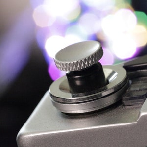 May include: A silver Canon QL17 camera with a black lens and a silver knob on top. The camera has a black leather strap attached to it.