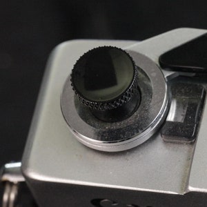 May include: A silver Canon Auto camera with a black knob on the top. The camera has a black leather strap attached to it.