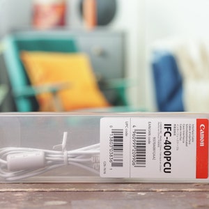 Puede incluir: Un cable de interfaz Canon IFC-400PCU blanco en un paquete de plástico transparente con una etiqueta roja y blanca. La etiqueta incluye el nombre del producto, el código EAN/JAN, el código UPC y el texto "Interface Cable, Cavo Interfaccia, Cable, インターフェースケーブル".