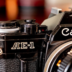 May include: A black Canon AE-1 film camera with a Canon FD 50mm f/1.8 lens. The camera has a black leather strap attached to it.