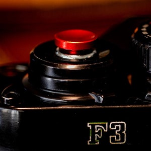 Puede incluir: Una cámara de película Nikon F3 negra con un botón rojo y una perilla plateada. La cámara tiene una inscripción plateada que dice "F3".