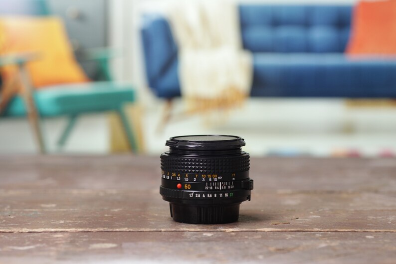 May include: A black camera lens with a silver ring and markings. The lens is sitting on a wooden surface.