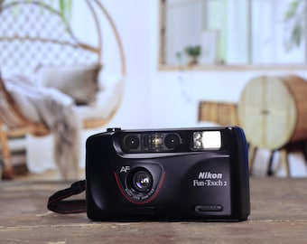 May include: Black Nikon Fun-Touch 2 camera with a red accent around the lens. The camera has a built-in flash and a wrist strap. The text "Nikon Fun-Touch 2" is printed on the front of the camera.