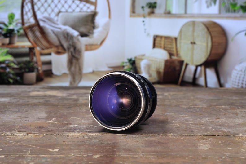May include: A close-up of a camera lens with a black and silver finish. The lens has a reflective, purple-tinted glass. The lens is resting on a weathered, brown wooden surface. The background is blurred, showing a room with a hanging chair.
