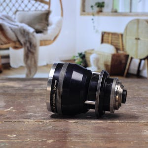 May include: A black camera lens with silver detailing, resting on a wooden surface. The lens features rings and markings, including "f=36mm...82mm". The background is blurred, revealing a room with a hanging chair and decorative elements.