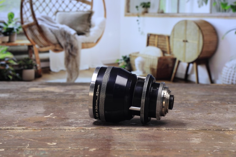 May include: A black camera lens with silver detailing, resting on a wooden surface. The lens features rings and markings, including "f=36mm...82mm". The background is blurred, revealing a room with a hanging chair and decorative elements.
