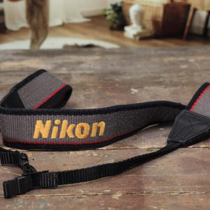 Può includere: Una tracolla per fotocamera Nikon grigia e nera con il nome del marchio ricamato in giallo. La tracolla ha linee di accento rosse e connettori in pelle e plastica neri. La tracolla è progettata per essere indossata intorno al collo.