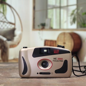 May include: A gold and black HC 2000 Focus Free camera. The camera has a red circle around the lens and the text "HC 2000 FOCUS FREE" on the front. The camera is sitting on a wooden surface.