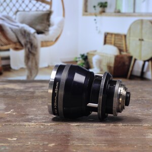 May include: A black camera lens with silver detailing, positioned on a wooden surface. The lens features a cylindrical design with rings and markings, including "f=36mm...82mm". The background is blurred, indicating an indoor setting.