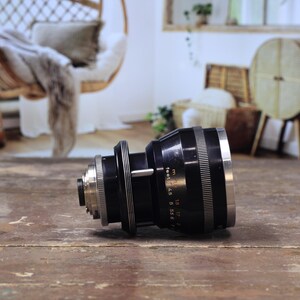 May include: A black and silver camera lens with measurement markings, resting on a wooden surface. The lens features a series of rings and a silver front element. The background is blurred, showing a room with a hanging chair.