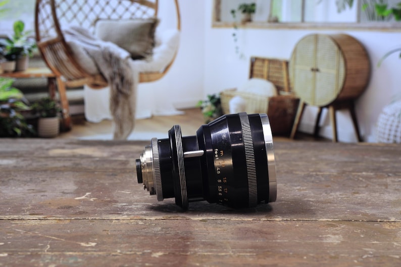 May include: A black and silver camera lens with measurement markings, resting on a wooden surface. The lens features a series of rings and a silver front element. The background is blurred, showing a room with a hanging chair.