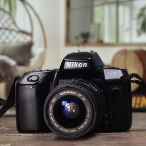 May include: A black Nikon N70 film camera with a Quantaray lens, on a wooden surface. The camera strap is visible. The background includes a hanging chair and a window.