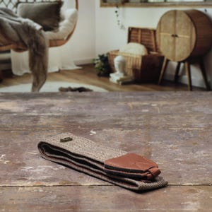 May include: A folded brown and tan herringbone guitar strap with a brown leather end piece and a metal buckle. The strap is lying on a weathered wooden surface.