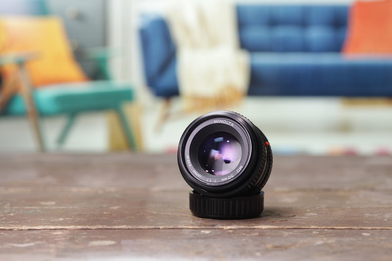 May include: A black Minolta MD 50mm f/1.7 lens with a silver ring around the lens barrel. The lens is sitting on a wooden table.