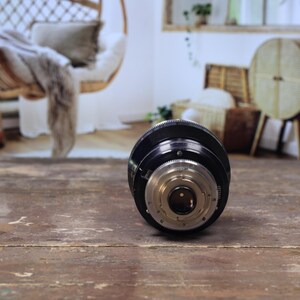 May include: A black and silver camera lens, positioned on a distressed wooden surface. The lens features intricate detailing with various rings and a central aperture. The background is blurred, hinting at a room with natural light and decorative items.
