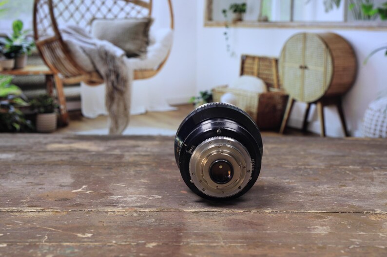 May include: A black and silver camera lens, positioned on a distressed wooden surface. The lens features intricate detailing with various rings and a central aperture. The background is blurred, hinting at a room with natural light and decorative items.