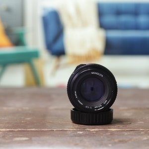 May include: A black Minolta MC 50mm 1:1.7 lens with a blue filter in the front. The lens is sitting on a wooden table.