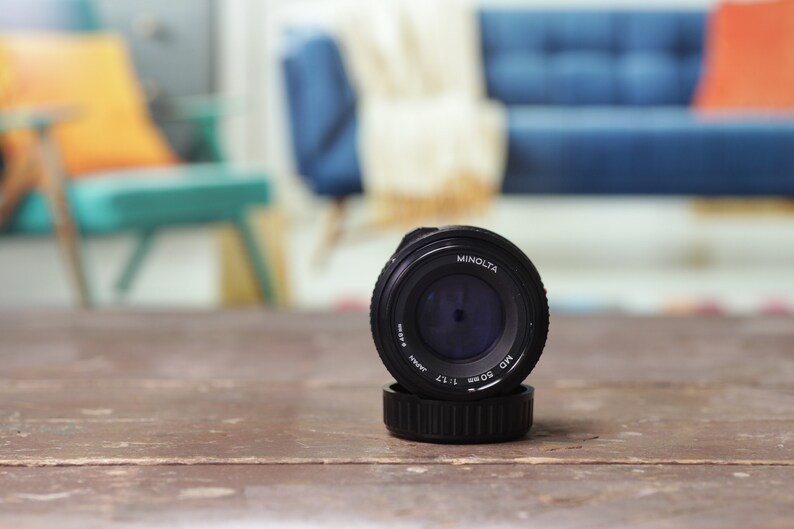 May include: A black Minolta MC 50mm 1:1.7 lens with a blue filter in the front. The lens is sitting on a wooden table.