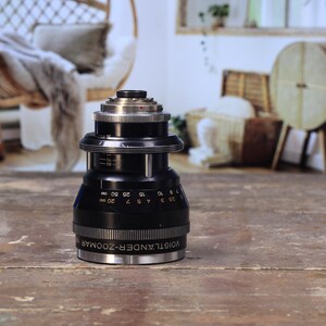 May include: A black and silver Voigtlander Zoomar camera lens. The lens has a series of numbers and markings, including the text "Voigtlander-Zoomar". The lens is cylindrical with a ribbed grip and sits on a wooden surface.