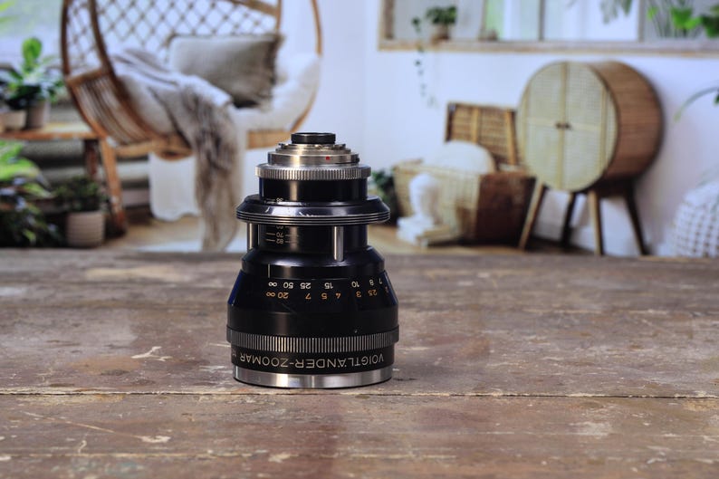 May include: A black and silver Voigtlander Zoomar camera lens. The lens has a series of numbers and markings, including the text "Voigtlander-Zoomar". The lens is cylindrical with a ribbed grip and sits on a wooden surface.