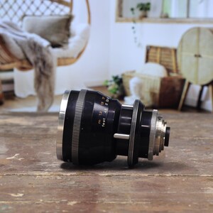 May include: A black and silver camera lens with measurement markings in feet. The lens is lying on a weathered wooden surface. The lens has a series of rings and a silver front element.