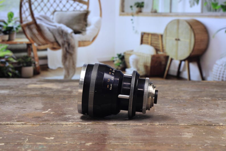 May include: A black and silver camera lens with measurement markings in feet. The lens is lying on a weathered wooden surface. The lens has a series of rings and a silver front element.