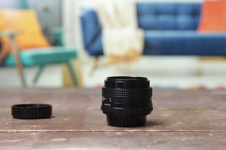 May include: A black camera lens with a black lens cap on a wooden table. The lens has a silver ring around it and a red dot on the side.