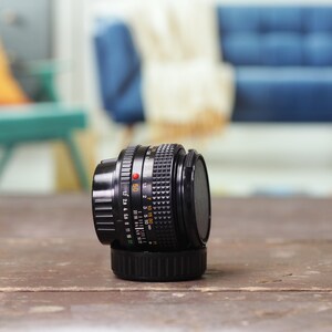 May include: A black camera lens with a silver ring and markings. The lens is sitting on a wooden surface.