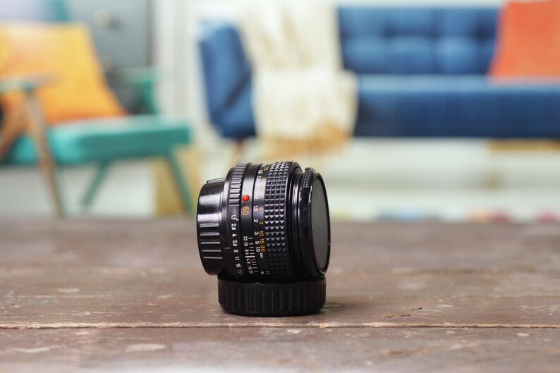 May include: A black camera lens with a silver ring and markings. The lens is sitting on a wooden surface.