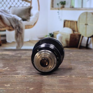 May include: A black camera lens with silver detailing lies on a worn wooden surface. The lens has a cylindrical shape with intricate internal components visible. The background shows a blurred interior with wicker furniture and plants.