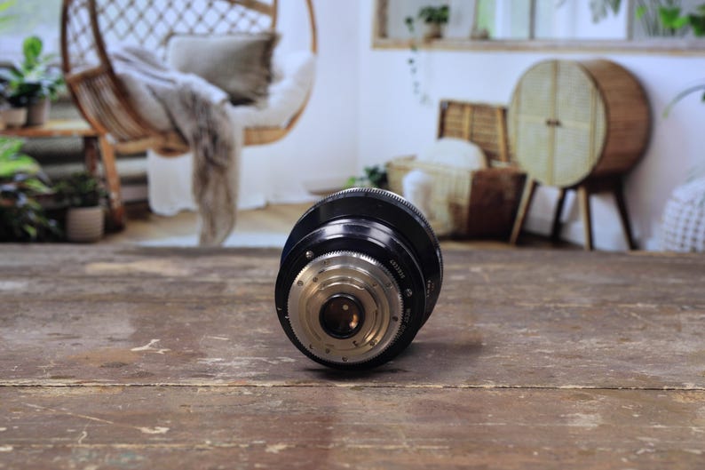 May include: A black camera lens with silver detailing lies on a worn wooden surface. The lens has a cylindrical shape with intricate internal components visible. The background shows a blurred interior with wicker furniture and plants.