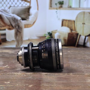 May include: A black and silver Voigtlander-Zoomar 1:2.8 lens, resting on a wooden surface. The lens features rings and markings, including distance scales. This is a piece of photography equipment.