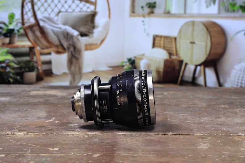 May include: A black and silver Voigtlander-Zoomar 1:2.8 lens, resting on a wooden surface. The lens features rings and markings, including distance scales. This is a piece of photography equipment.