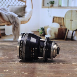 May include: A black and silver camera lens with the text "Schneider-Zoomar 2.8 f=28-85mm" printed on the side. The lens is lying on a wooden surface. The background includes a hanging chair and other decorative items.