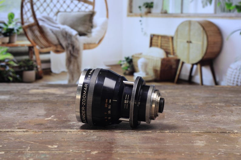 May include: A black and silver camera lens with the text "Schneider-Zoomar 2.8 f=28-85mm" printed on the side. The lens is lying on a wooden surface. The background includes a hanging chair and other decorative items.